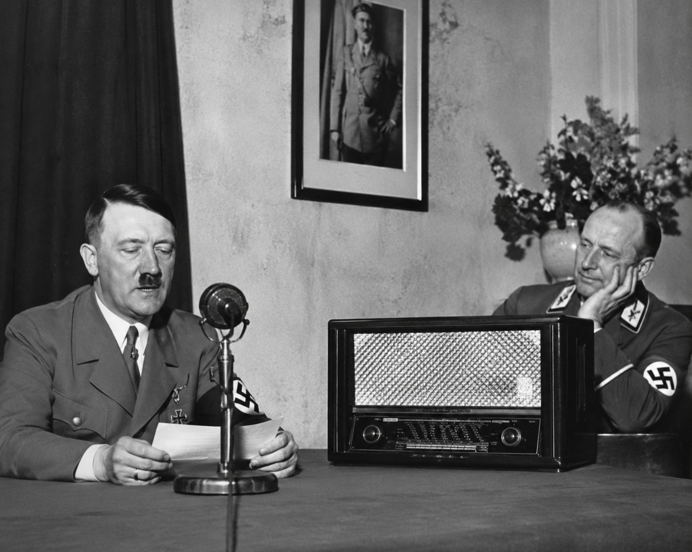 Adolf Hitler spreekt in een microfoon terwijl een minister van de nazipartij naast een Volksempfänger radio luistert.